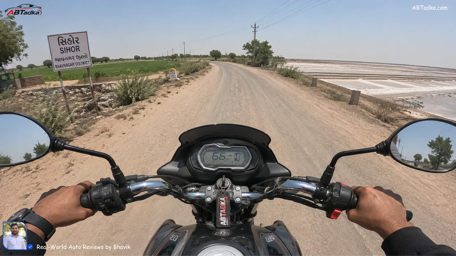 Bhavik Munjapara from ABTADKA.com testing a 125cc bike on the dusty village road outside Sihor, Bhavnagar, with farms and salt pans visible in the background during a hot Gujarat afternoon - 125cc vs 100cc