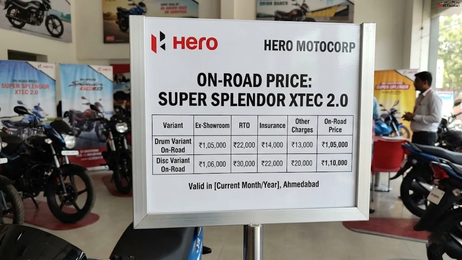 A Hero MotoCorp showroom on-road price board for the Super Splendor XTEC 2.0 in Ahmedabad, showing a comparison table of ex-showroom prices, RTO, insurance, and other charges for both Drum and Disc variants.