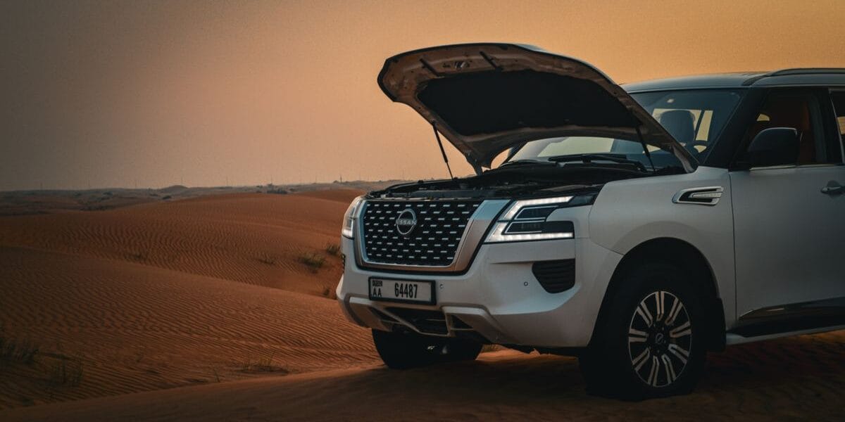 A white SUV with an open hood parked on scenic desert dunes during sunset.